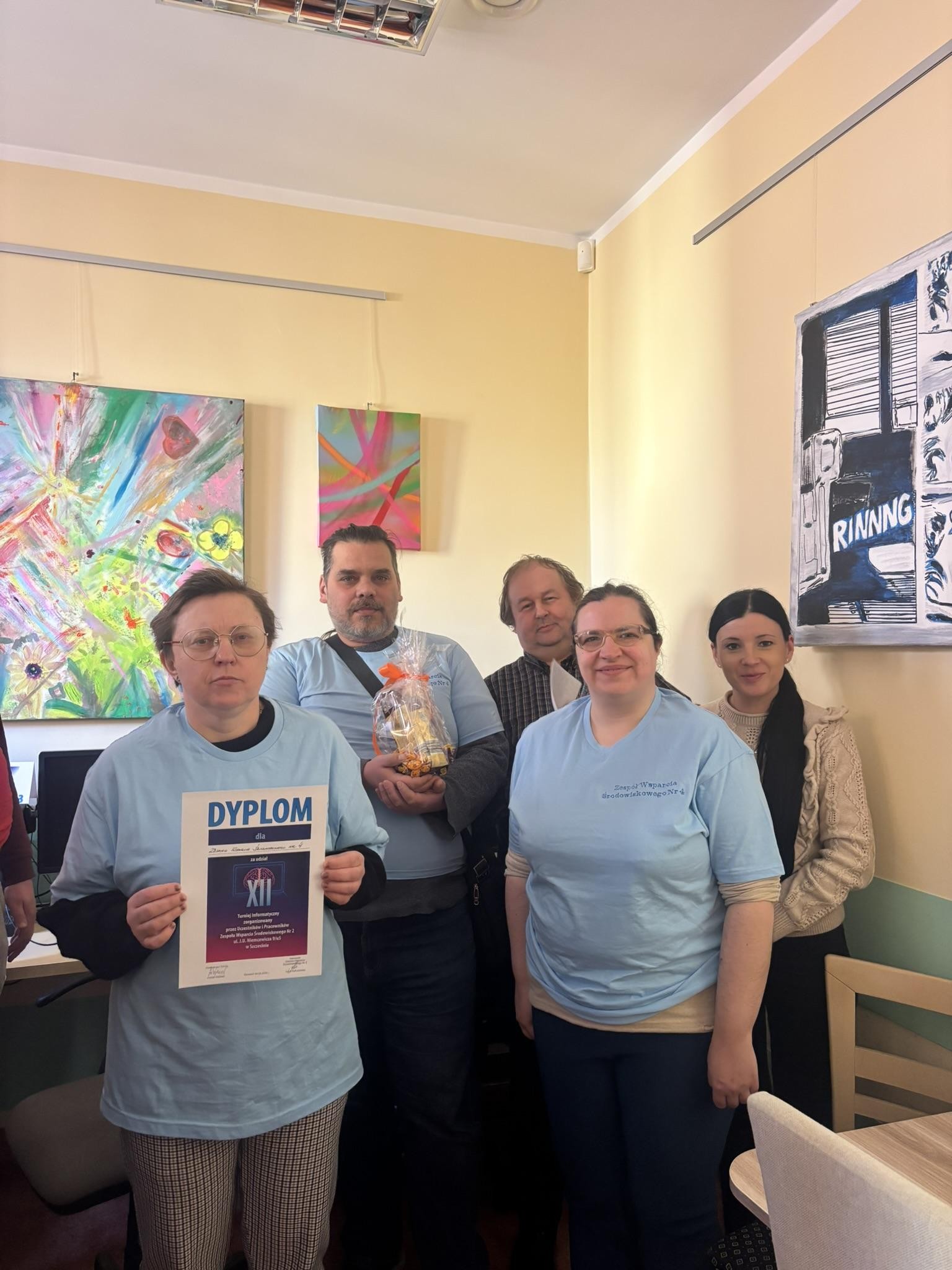 Grupa pięciu osób. Trzy osoby stojące na pierwszym planie mają na sobie niebieskie koszulki. W tle widać trzy obrazy wiszące na ścianach. 