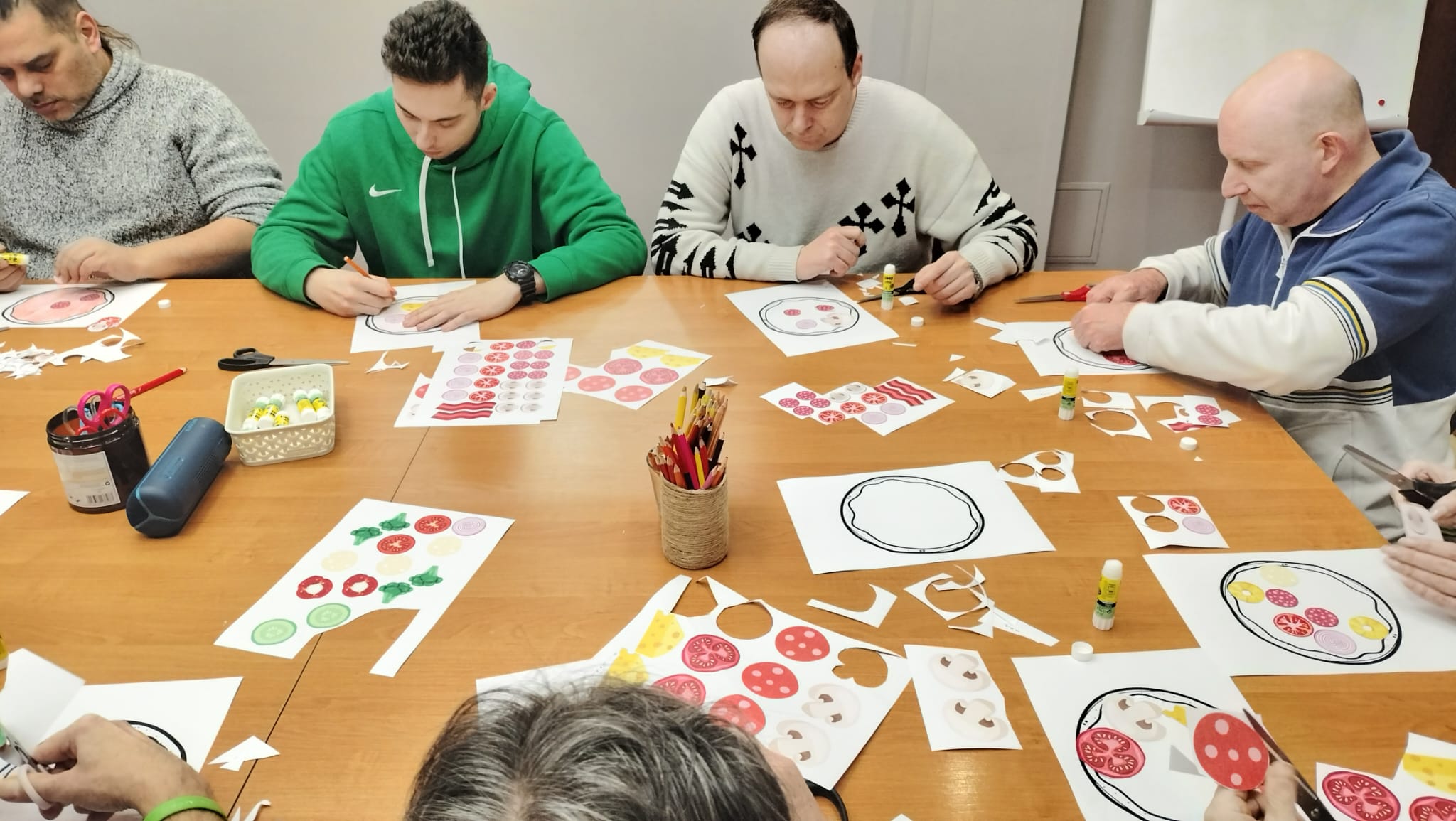 Grupa osób siedząca przy stole. Osoby wycinają kolorowe elementy z kartek. 