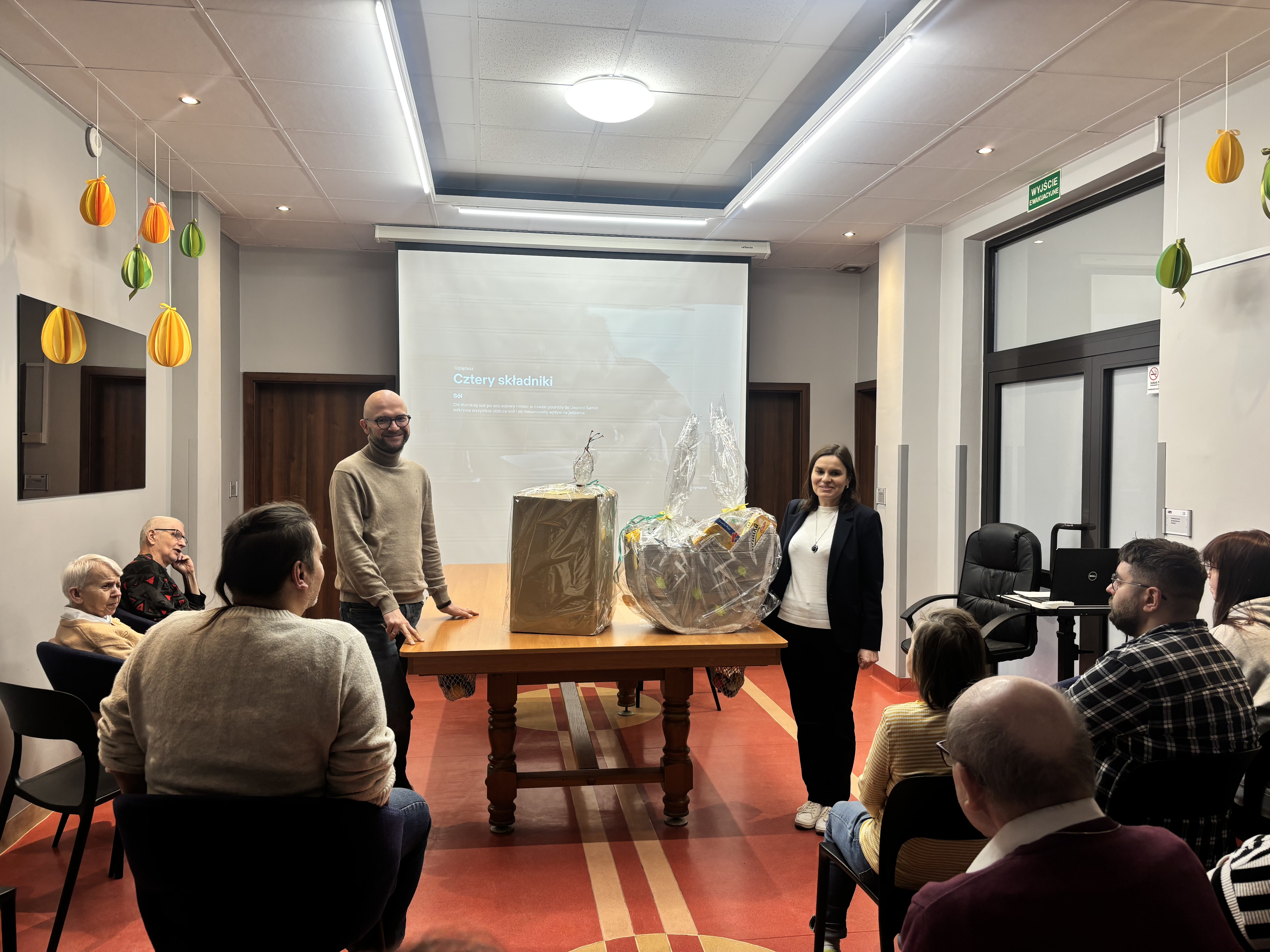 Na środku zdjęcia są dwie stojące osoby. Pomiędzy nim duży stół. Na stole stoją dwa duże pakunki. Dookoła stołu siedzą ludzie na krzesłach. W tle widać ekran i drzwi.
