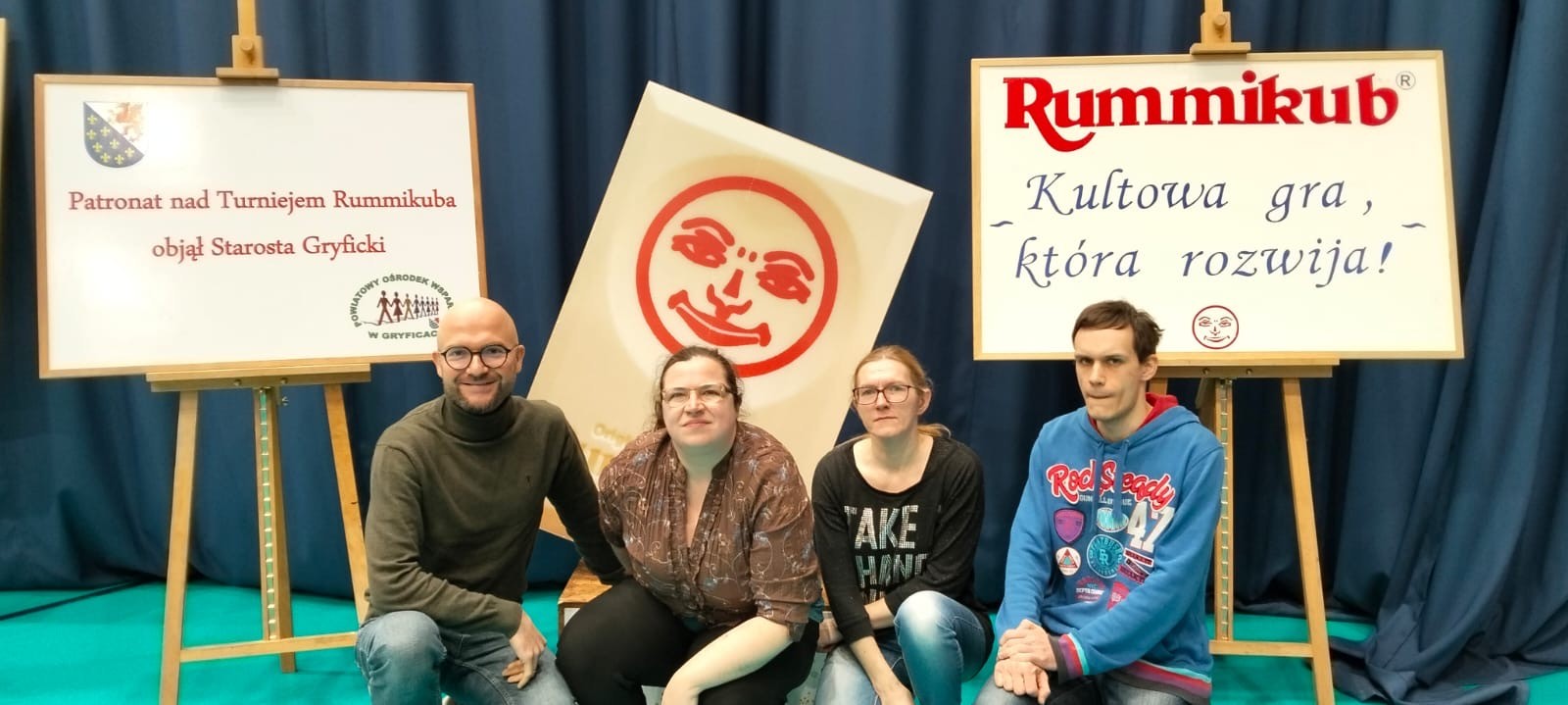 : Grupa czterech osób. Osoby kucają. W tle widać dwie tablice oraz dekorację. Na tablicy widać napis Rummikub oraz logo gry.