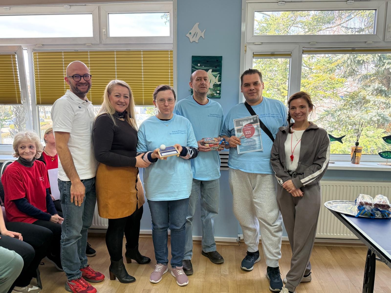 : Na pierwszym planie widoczna grupa sześciu stojących osób. Trzy z nich mają niebieskie koszulki oraz trzymają w rękach przedmioty. Na drugim planie widać siedzące na krzesłach osoby. W tle znajdują się dwa okna.