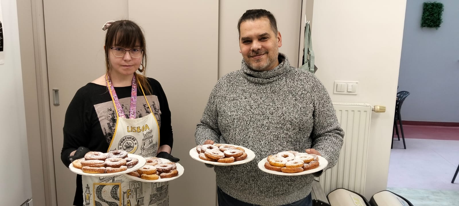 Kobieta i mężczyzna. Oboje trzymają białe talerze. Na talerzach oponki z cukrem pudrem. W tle zabudowa meblowa.