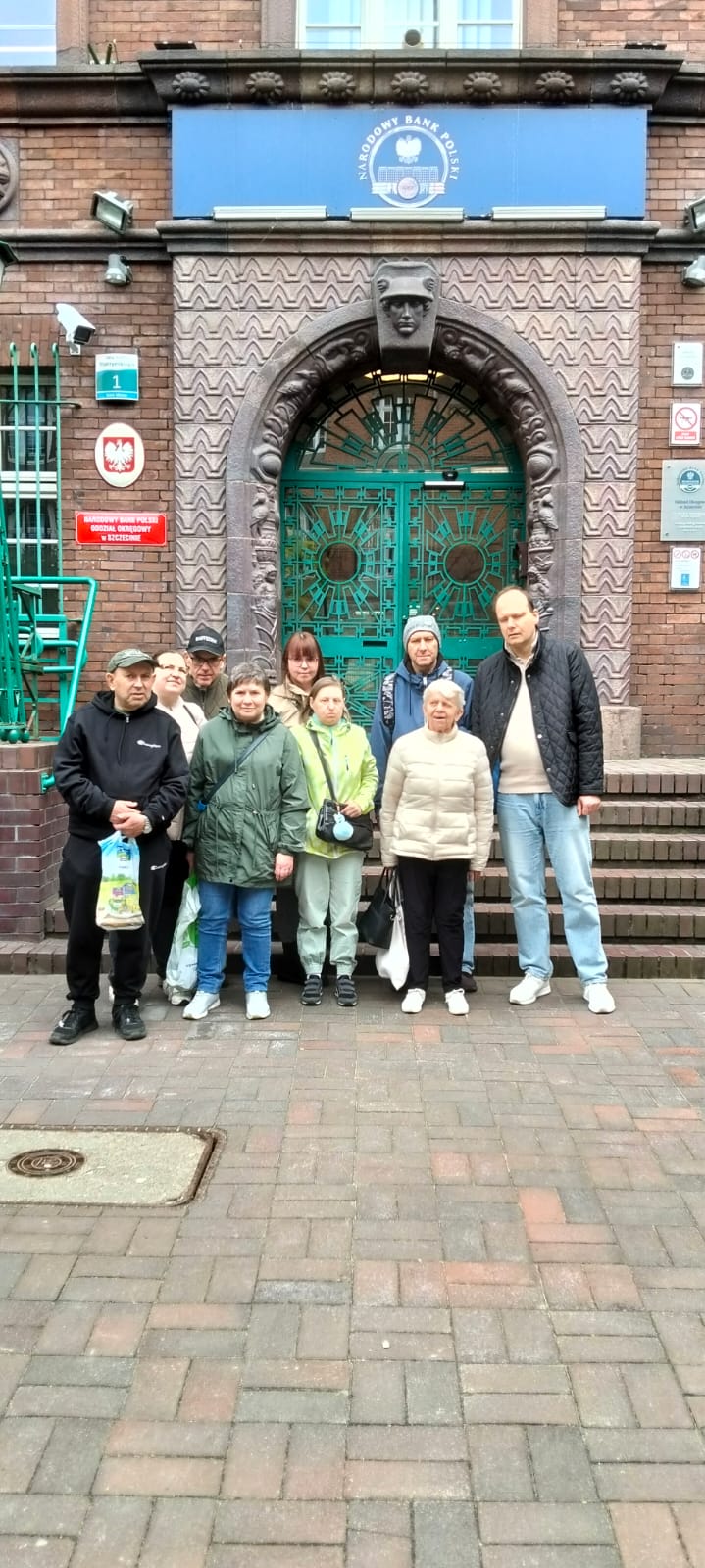 Grupa osób stojąca na tle budynku. Budynek jest w kolorze brązowym, drzwi są zielone. Jest na nim niebieski szyld.