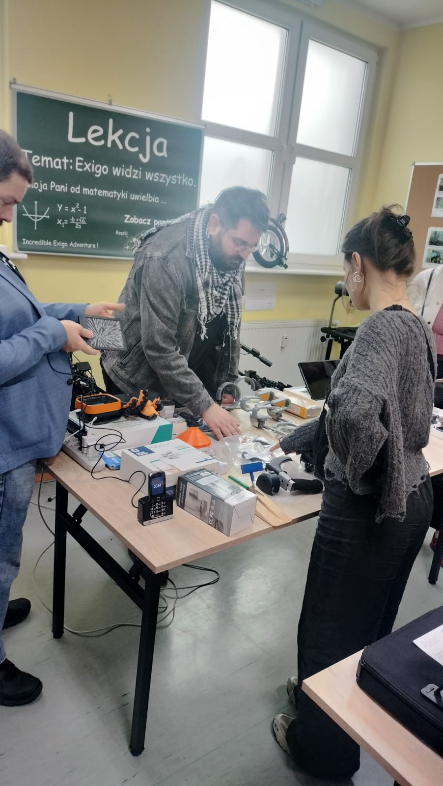 Grupa trzech osób. Trzy osoby stoją dookoła stołu. Na stole znajdują się różne przedmioty. W tle widać okno oraz szkolną tablice.