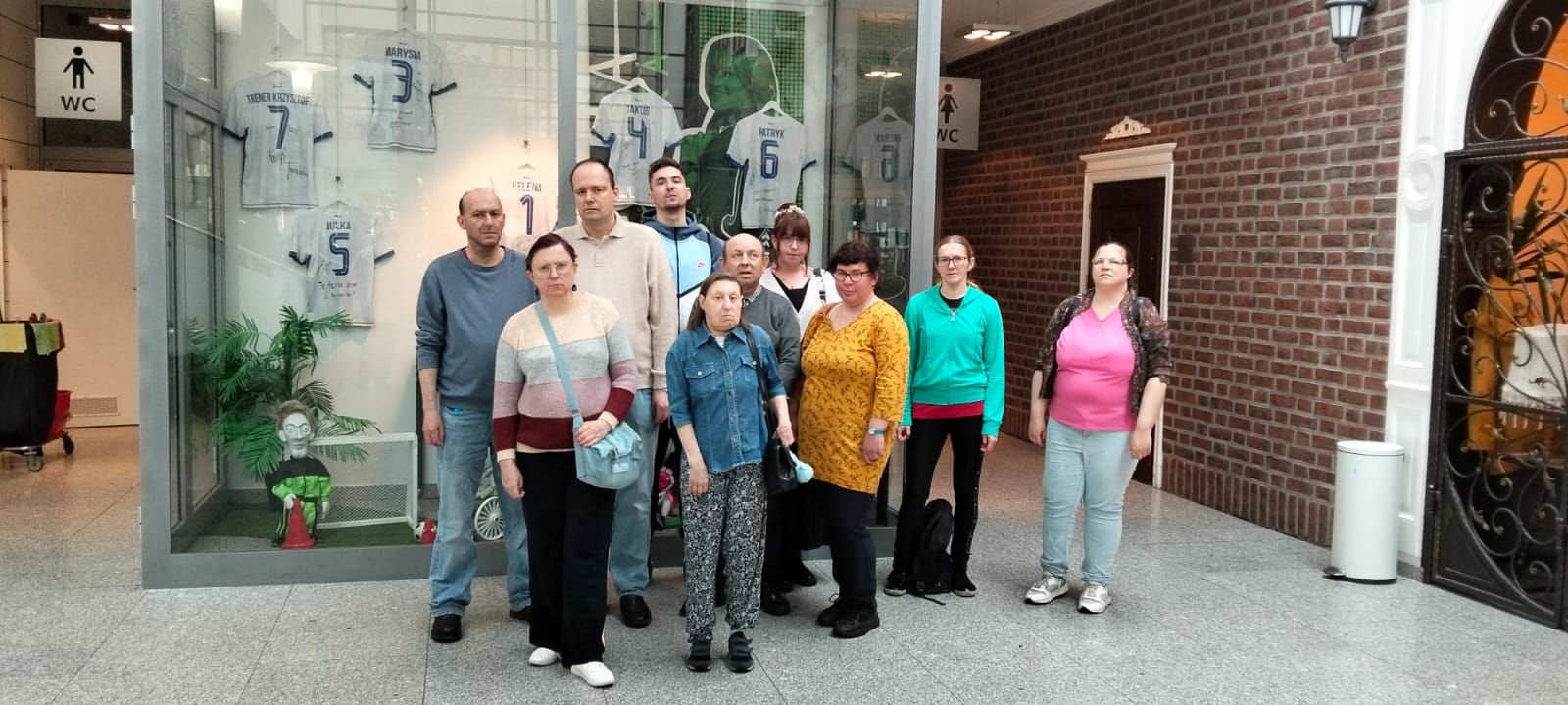Grupa stojących osób. W tle widać witrynę, ceglaną ścianę, biały kosz na śmieci oraz drzwi. W witrynie znajdują się koszulki sportowe.