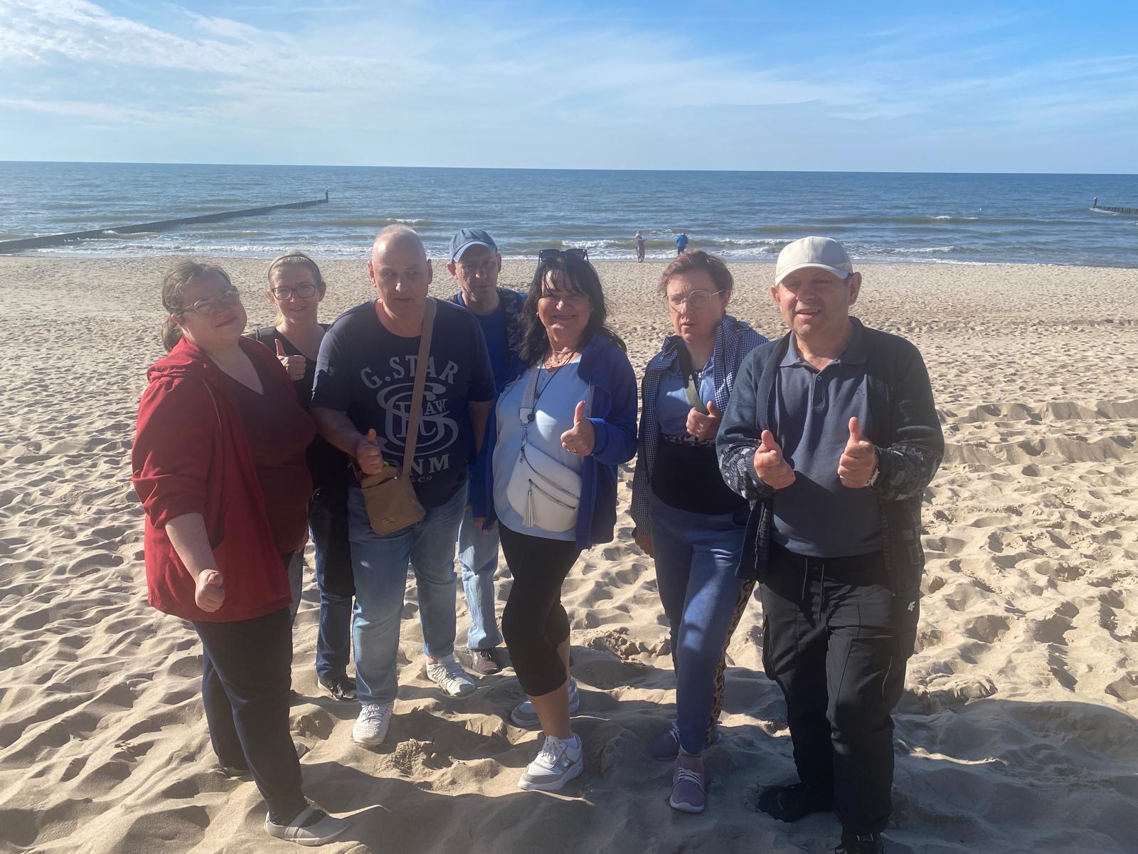 Grupa siedmiu osób stojąca na plaży. W tle widać morze.