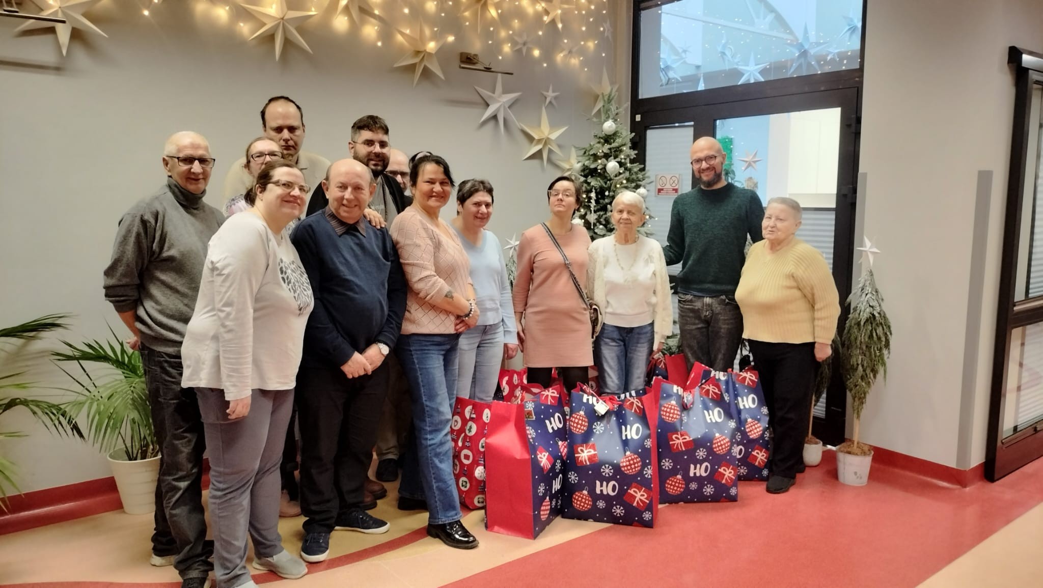 Grupa osób pozująca z dużymi torbami prezentowymi na tle dekoracji świątecznej.