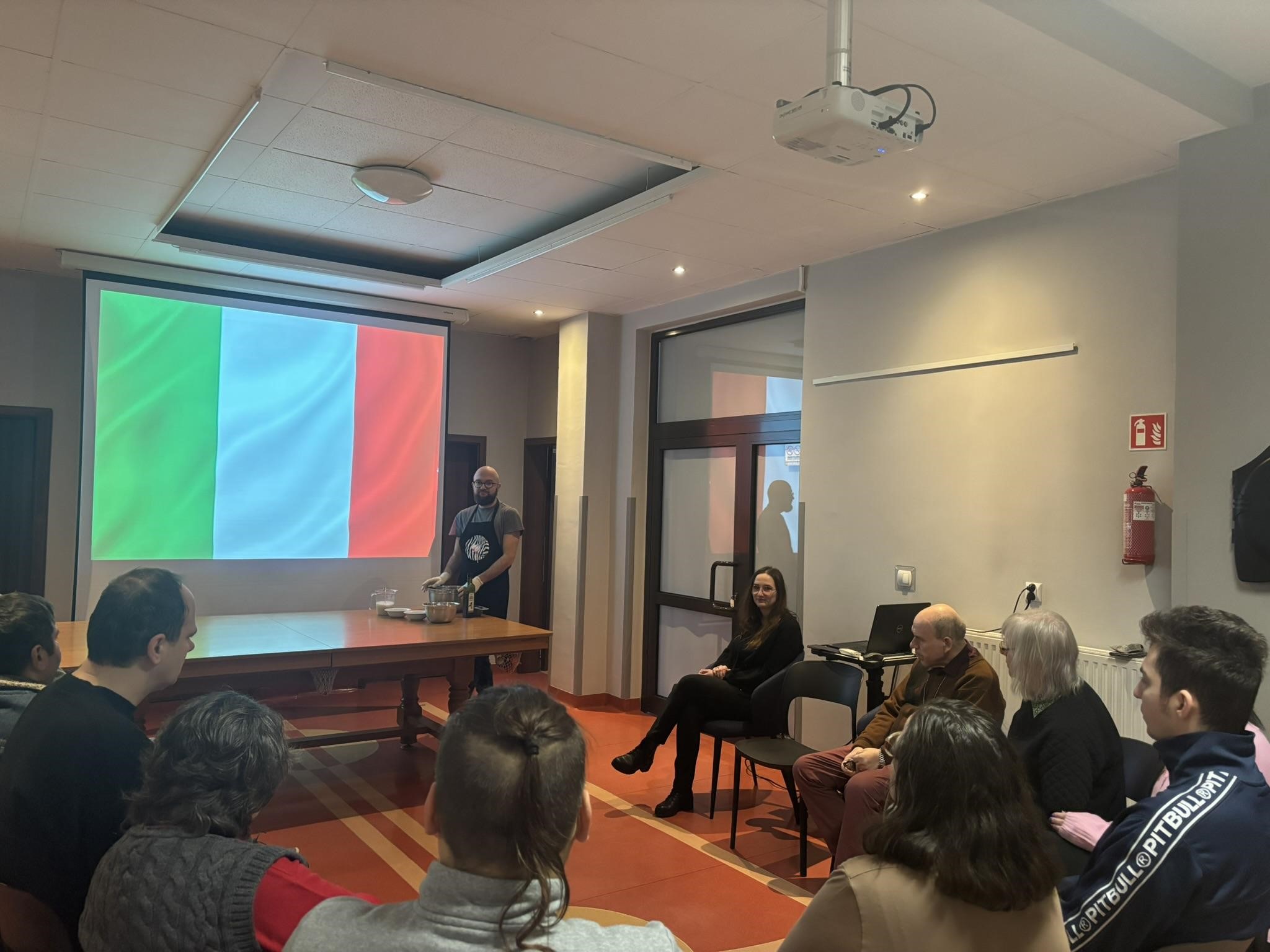 Grupa osób siedzi w półokręgu na krzesełkach. Przed nimi stoi mężczyzna. W tle widać wyświetloną na ekranie flagę Włoch.