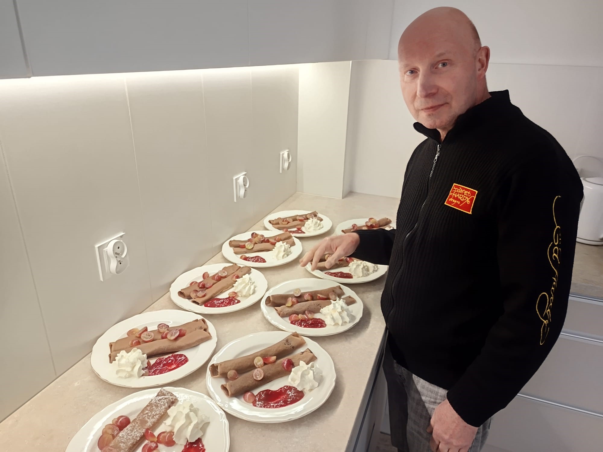 Mężczyzna stojący przy blacie kuchennym. Na blacie stoją talerze z naleśnikami, bitą śmietaną i owocami.