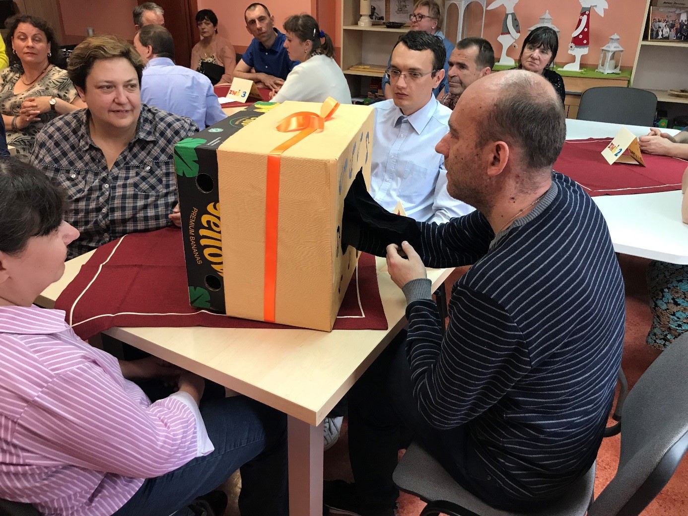 Grupa kobiet i mężczyzn rozpoznająca instrumenty ukryte w kartonie na podstawie dotyku. 