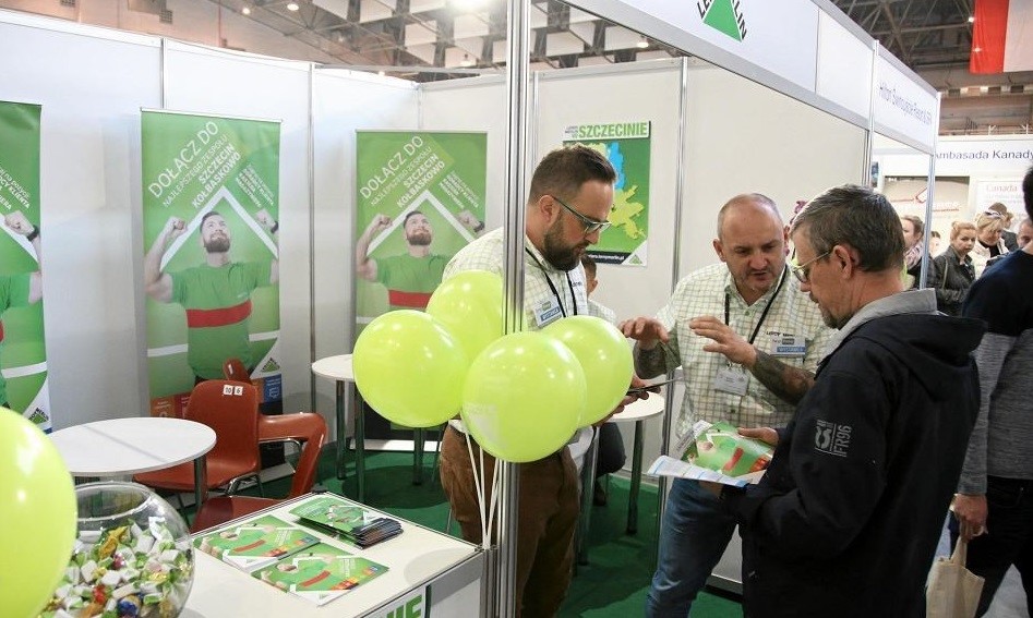 Trzech mężczyzn przy stanowisku prezentującym pracodawcę, w tle osoby odwiedzające targi pracy. 