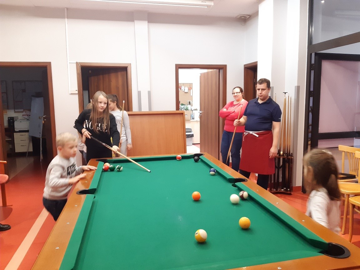 Grupa dzieci grających w bilard, w tle dwoje ludzi obserwujących grę. 