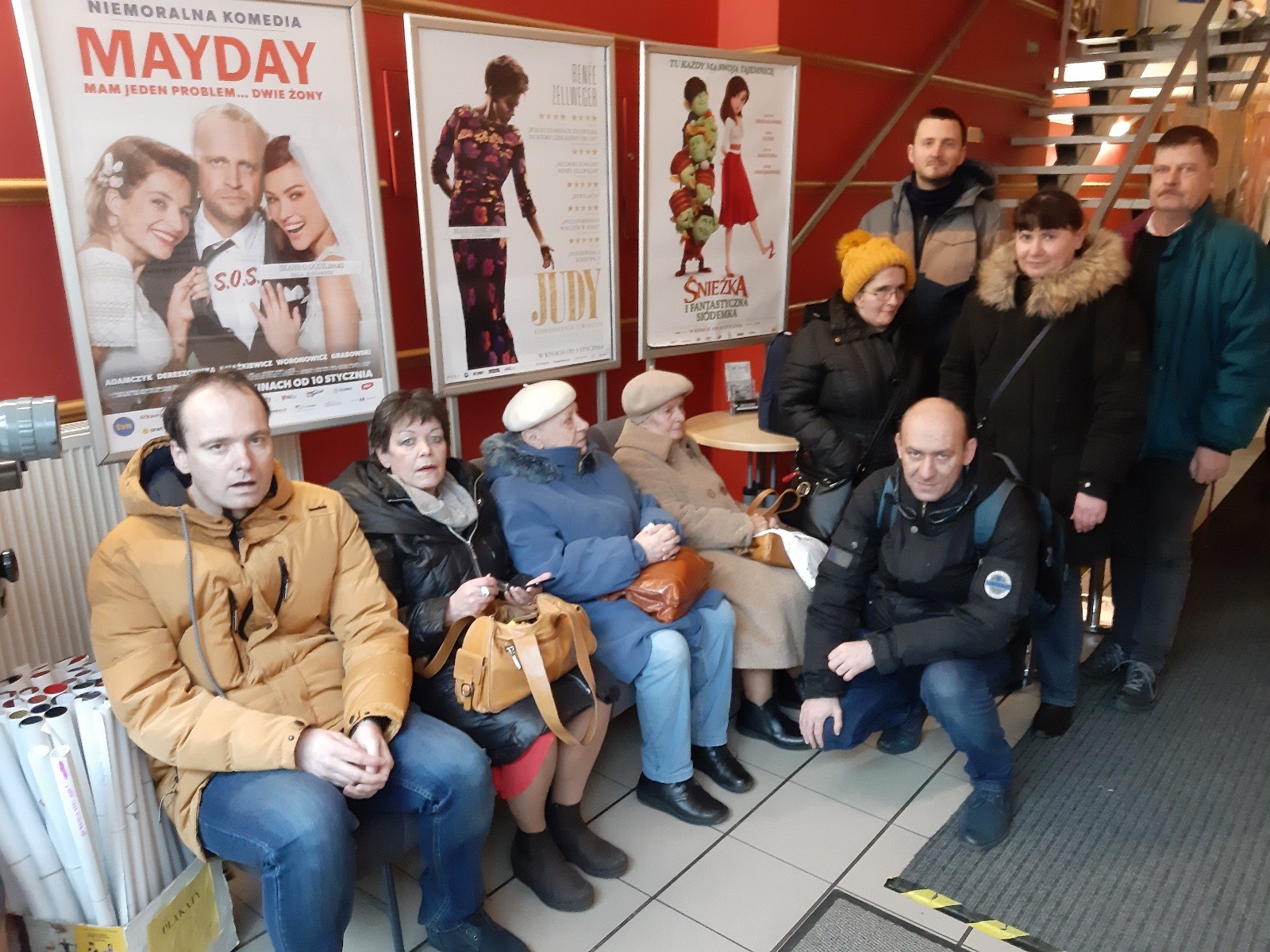 Grupa ludzi w oczekiwaniu na wejście do sali kinowej, w tle plakaty promujące filmy. 