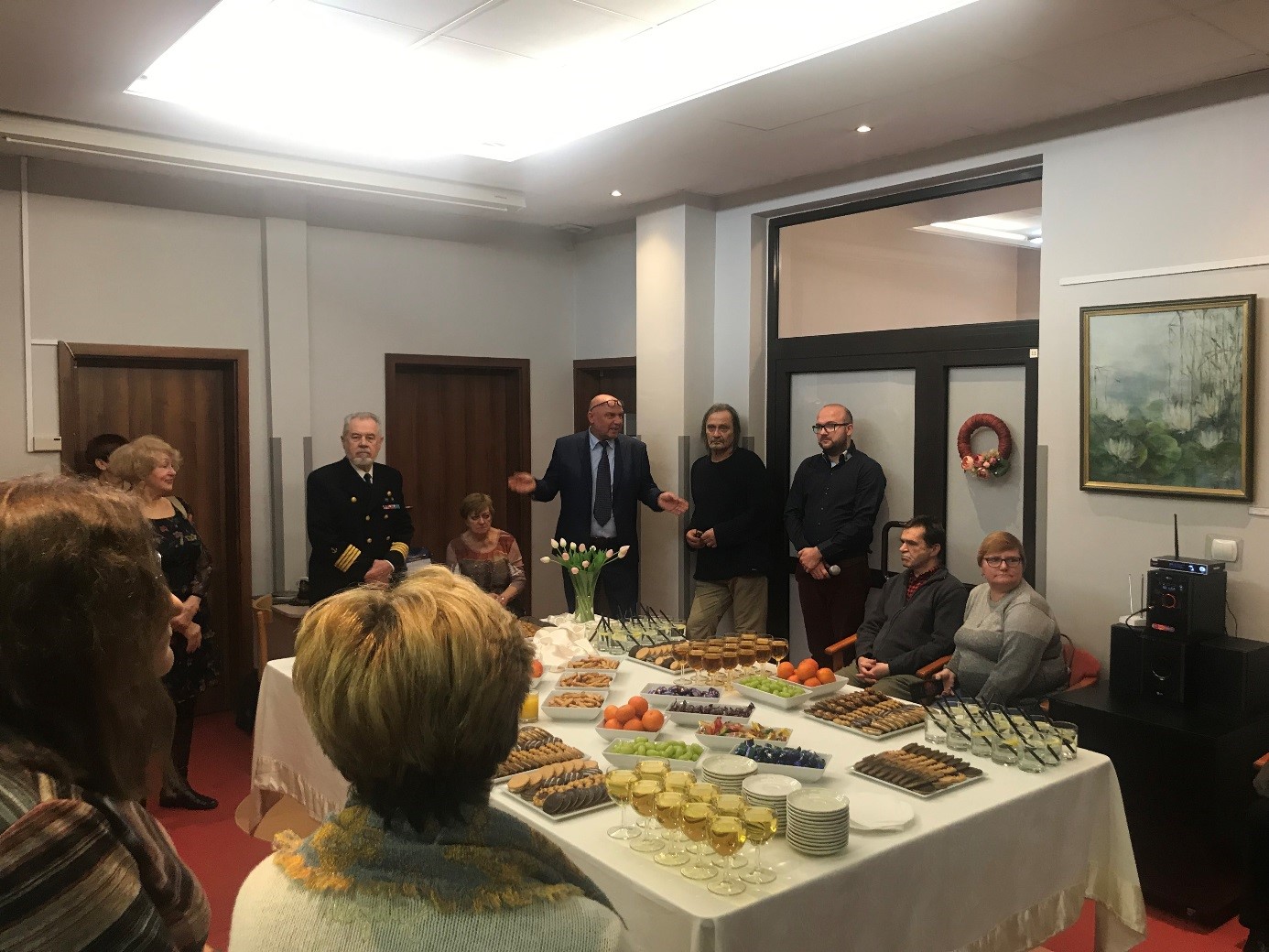 Grupa uczestników wernisażu podczas otwarcia imprezy zgromadzona wokół stołu z poczęstunkiem. 