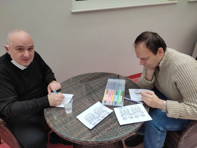 Dwaj mężczyźni przy stole kolorują elementy kart świątecznych.