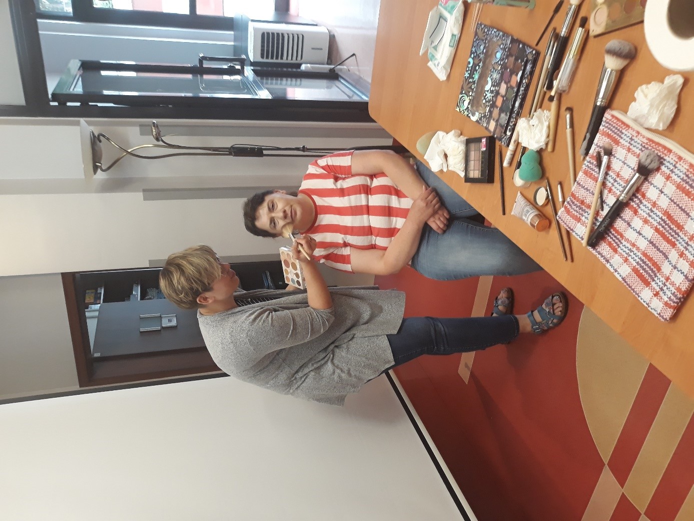 Dwie kobiety wykonujące makijaż dzienny. Na stole kosmetyki. 