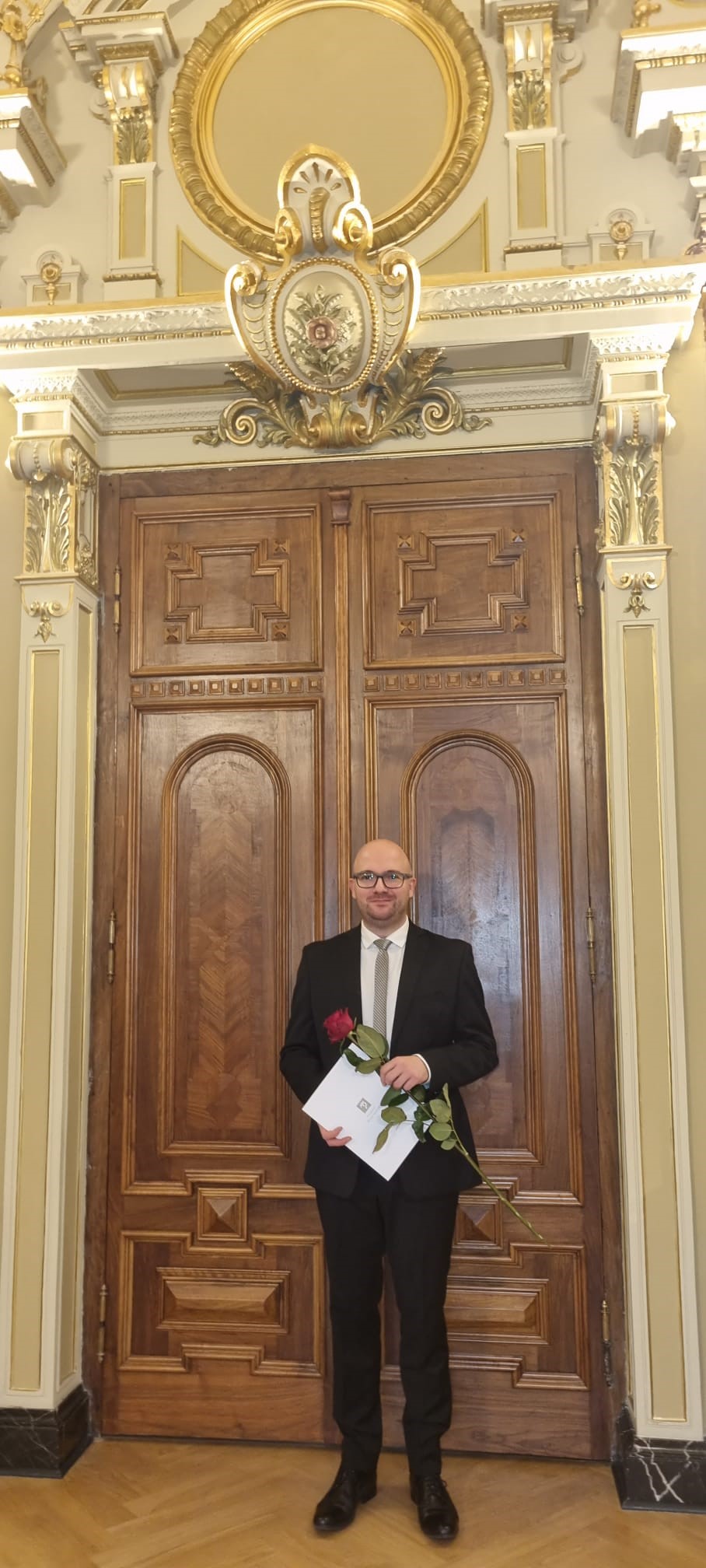 Mężczyzna w garniturze z dyplomem i różą. W tle zabytkowa architektura.