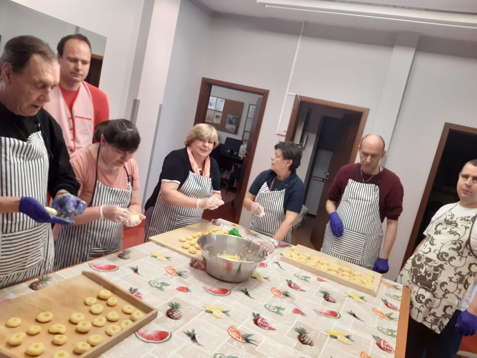 Grupa kobiet i mężczyzn w fartuchach kuchennych przy stole przygotowują kluski śląskie. 