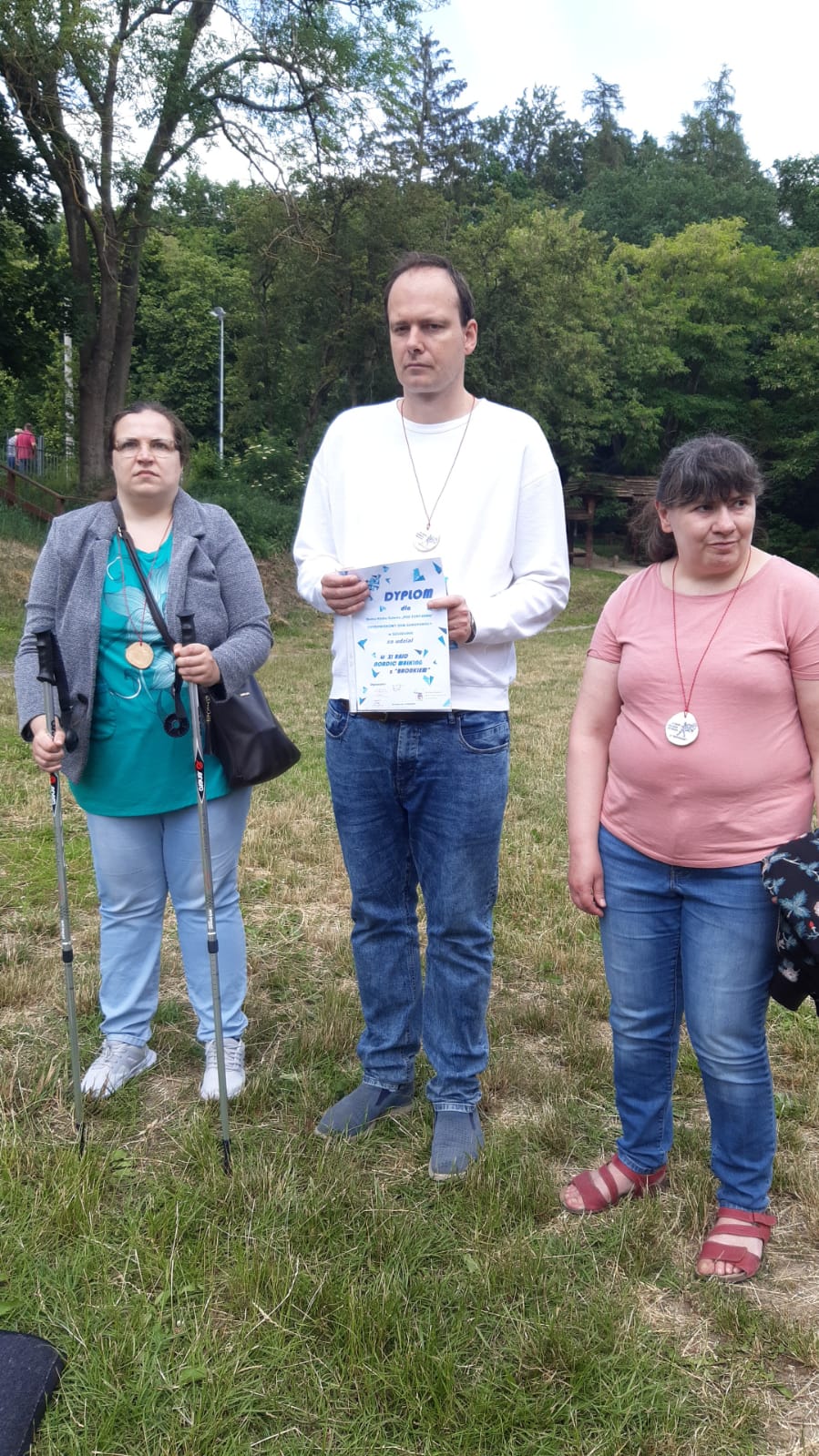 Dwie kobiety i mężczyzna na tle drzew, podczas wręczenia dyplomów i medali. 