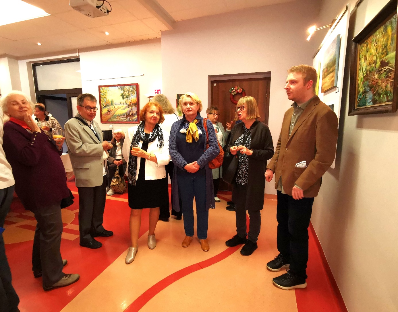 Grupa kobiet i mężczyzn podczas wernisażu, w tle obrazy. 