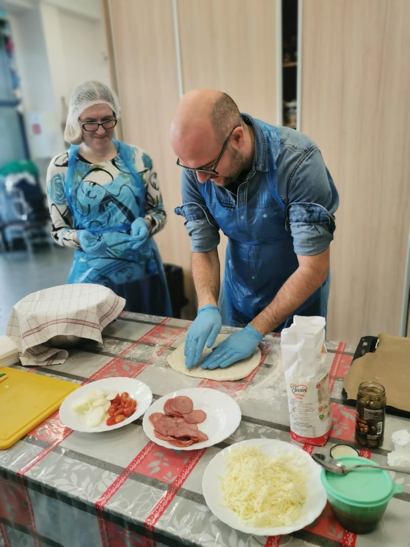Kobieta i mężczyzna przy stole podczas przygotowywania pizzy. Na pierwszym planie produkty spożywcze 
