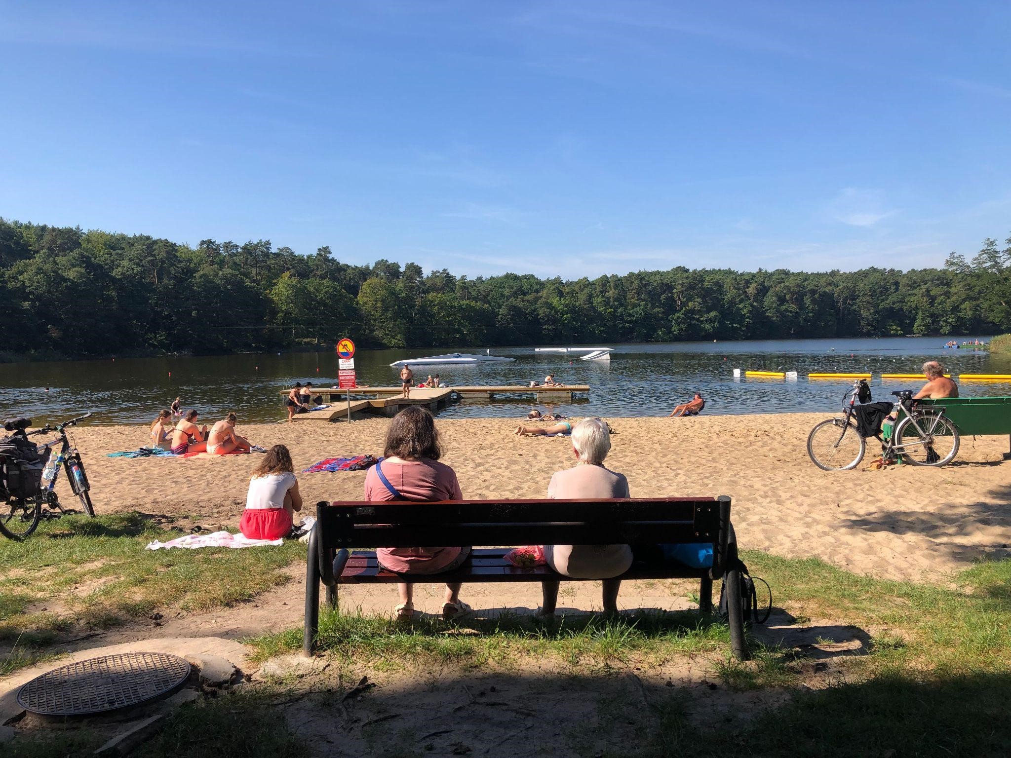Dwie zwrócone w stronę jeziora kobiety siedzące na ławce. W tle plażowicze.