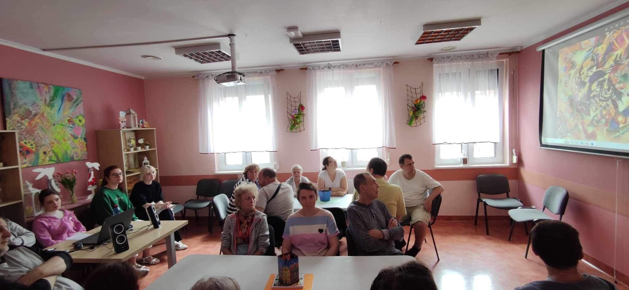 Grupa osób siedząca przy stołach podczas zajęć terapeutycznych.