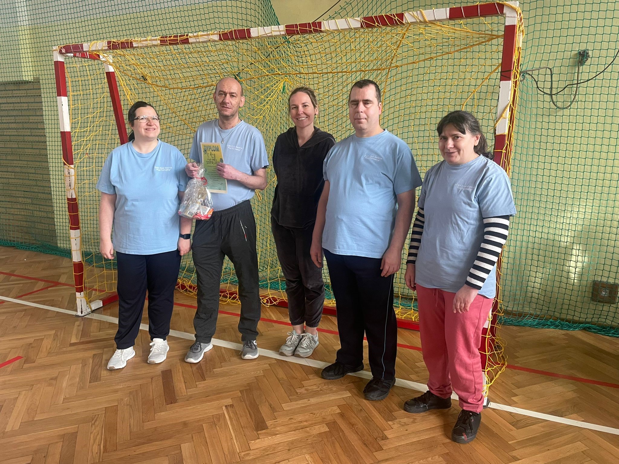 Grupa osób w strojach sportowych pozująca do zdjęcia na tle bramki piłkarskiej. 