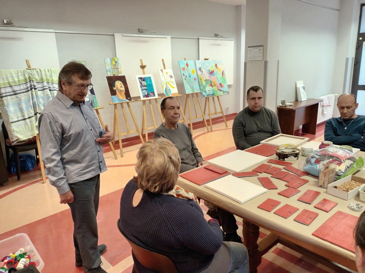 Grupa osób przy stole z artykułami plastycznymi. W tle sztalugi z obrazami. 