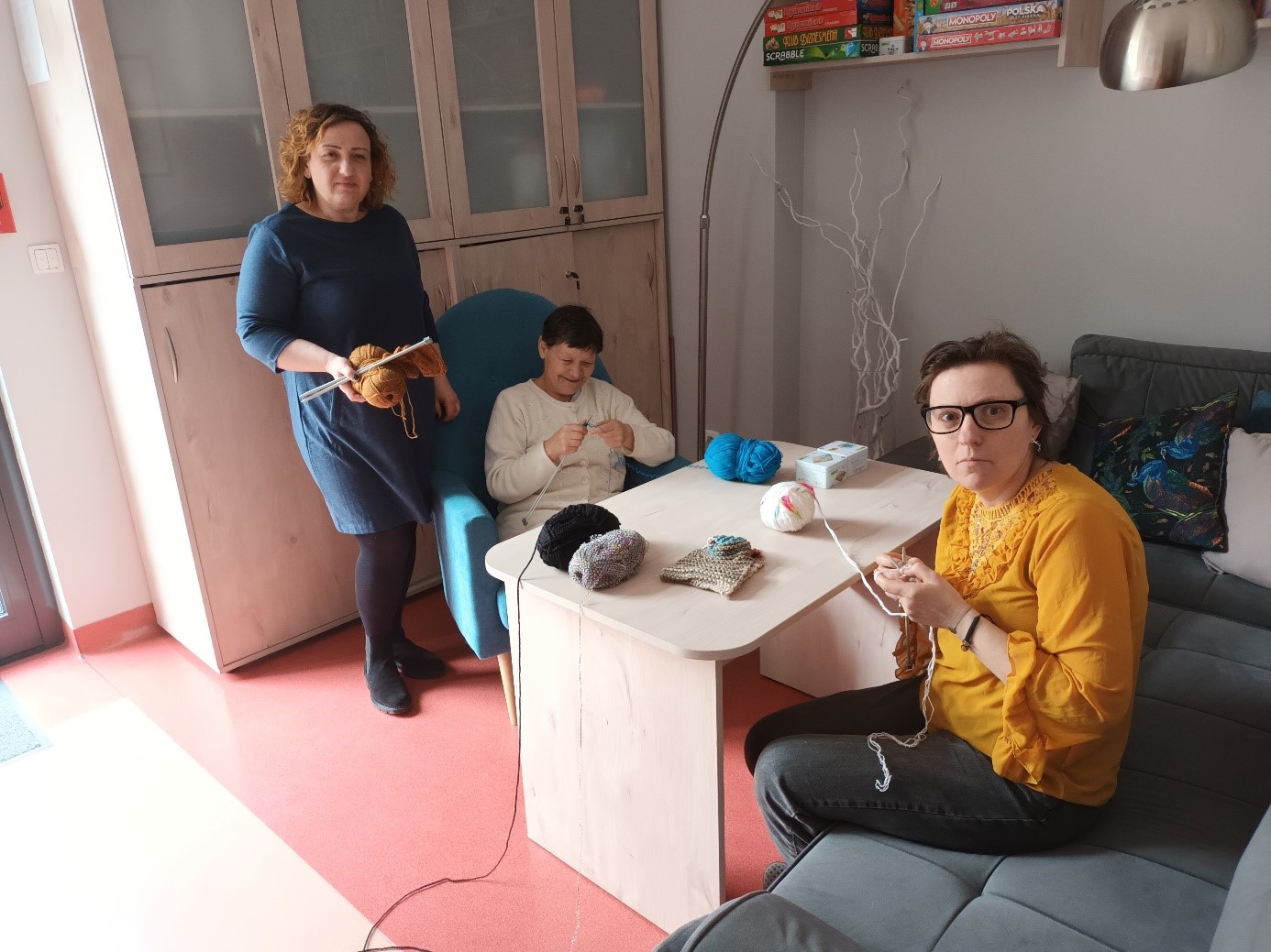 Trzy kobiety podczas robótek ręcznych na drutach. W tle zabudowa meblowa. 