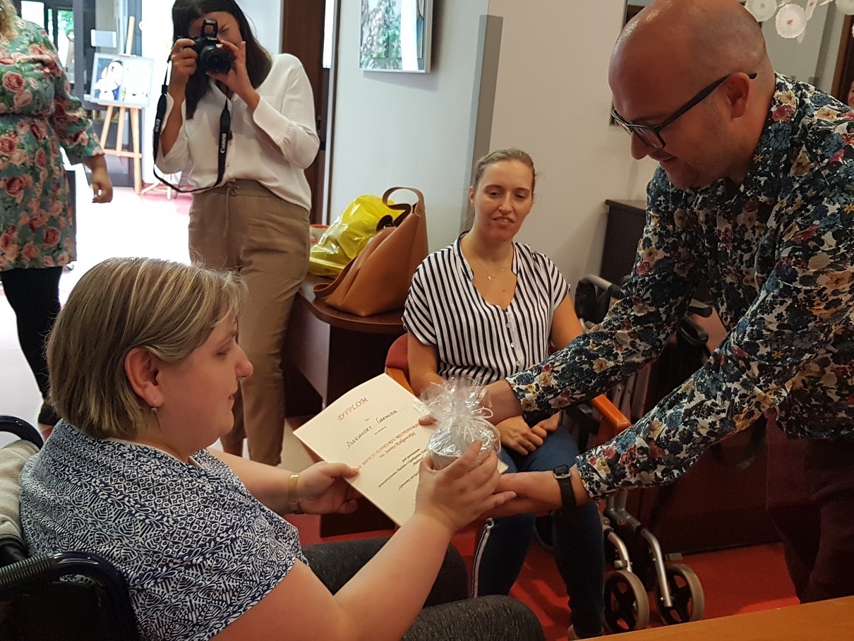 Terapeuta wręcza siedzącej uczestniczce dyplom i nagrodę. Za nimi siedzi uczestniczka. Obok niej stoi terapeutka, która trzyma w rękach aparat fotograficzny. 