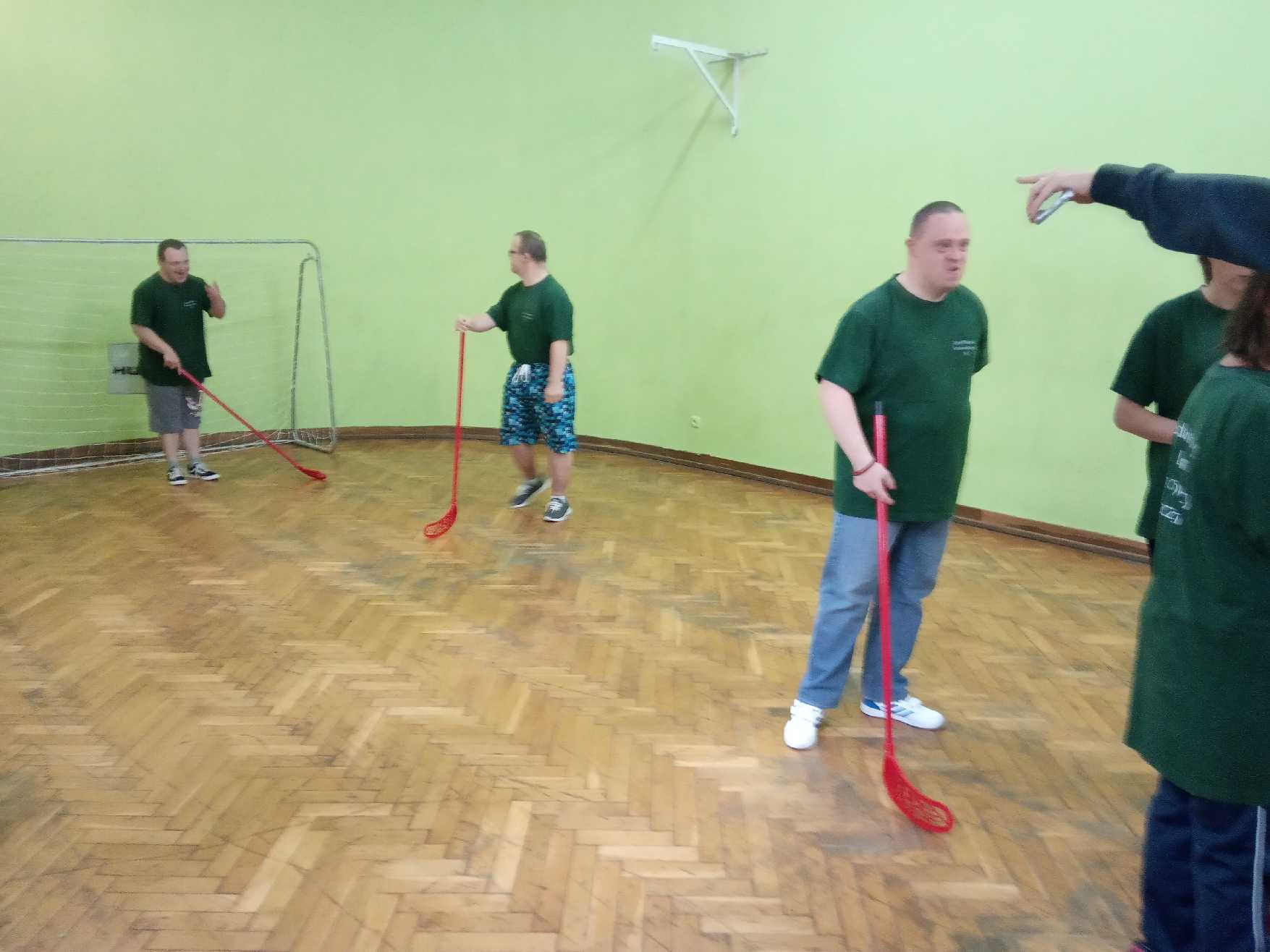 Troje uczestników stoi na hali sportowej. Każdy z nich w ręce trzyma kij do gry w hokeja. Jeden z nich stoi przy bramce. 