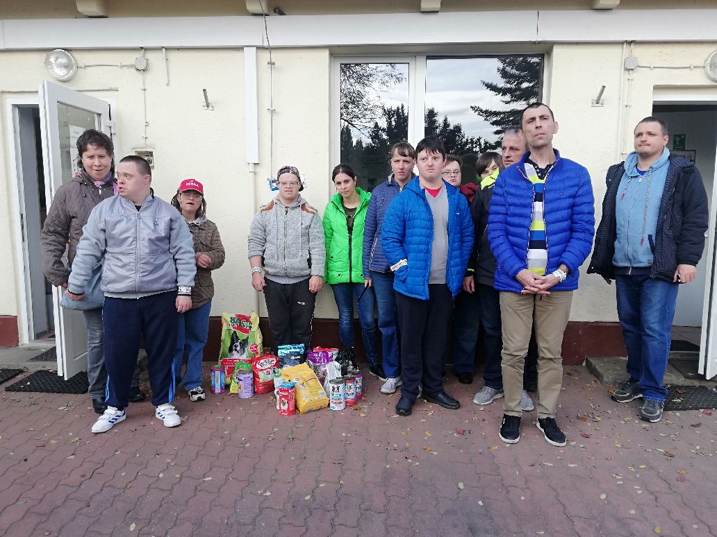 Przed budynkiem grupa osób stoi w rozsypce. Na ziemi przed grupą leżą opakowania z jedzeniem dla zwierząt.