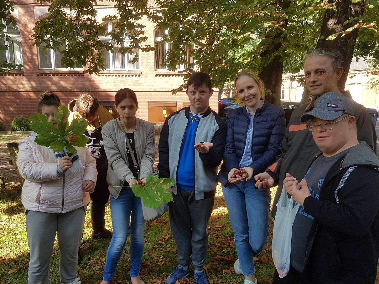 Grupa uczestników wraz z terapeutą stoją w półkolu pod drzewem. W rękach trzymają kasztany i liście. 