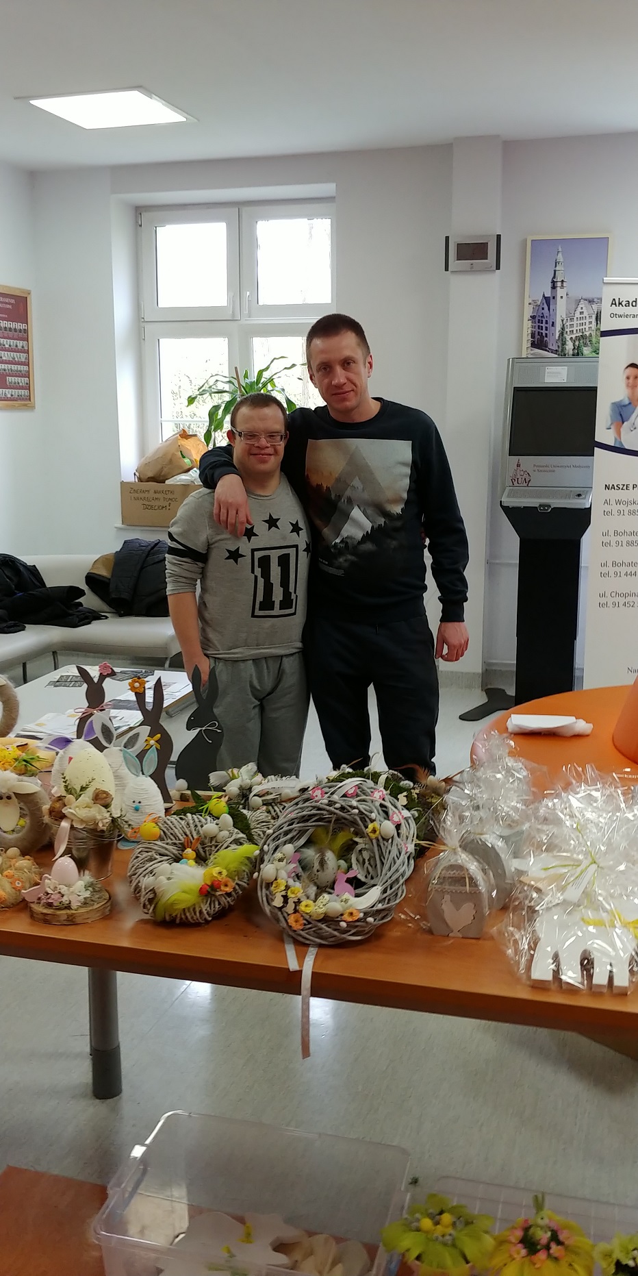 1.	Kiermasz świąteczny PUM  Terapeuta stoi obok uczestnika obejmując ręką jego ramiona. Przed nimi znajduje się stół z ozdobami wielkanocnymi. 