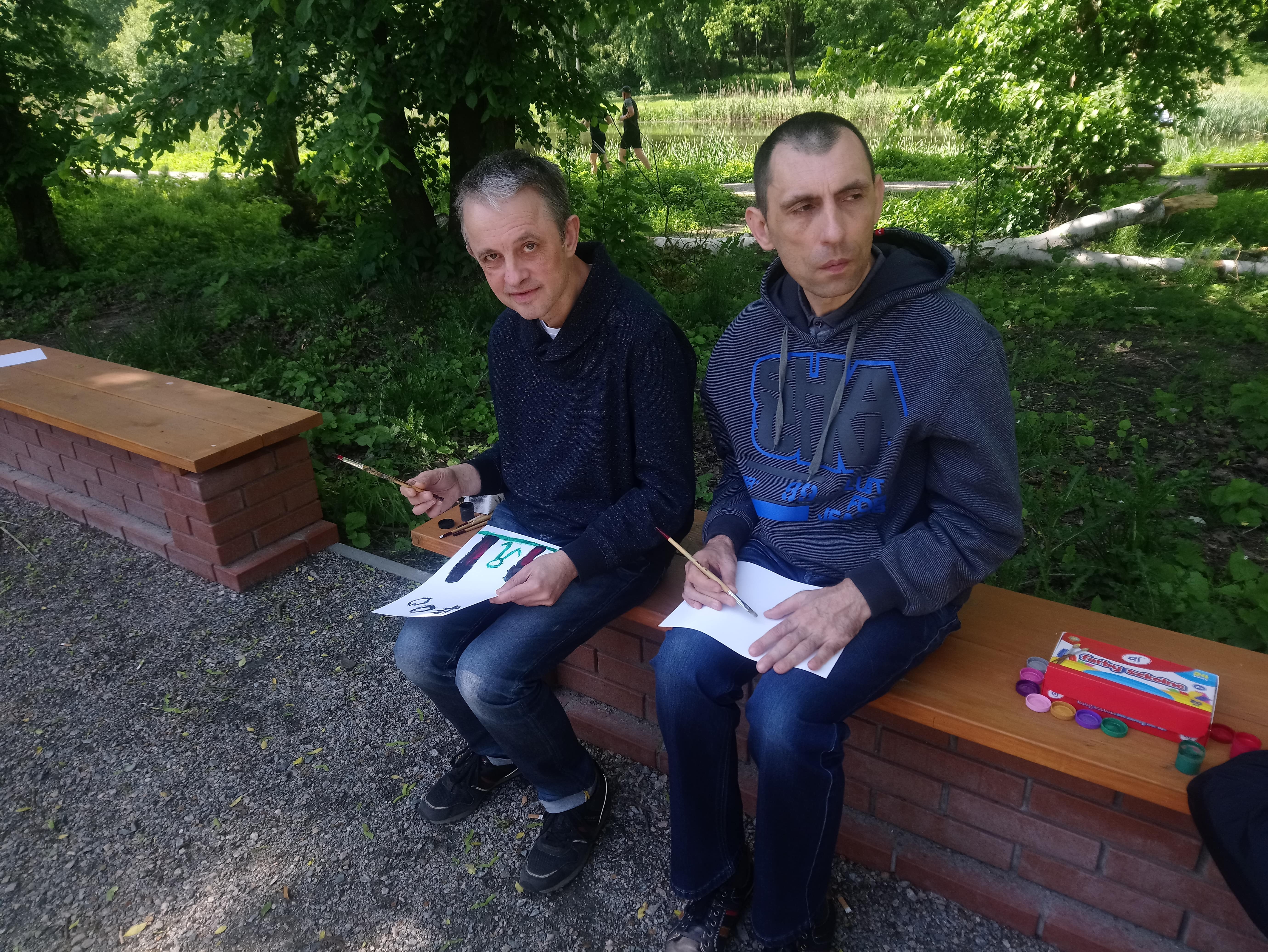 Dwóch uczestników siedzi na ławce w parku. Na kolanach trzymają kartki papieru, a w rękach pędzel. Na brzegu ławki leży opakowanie z farbami. 