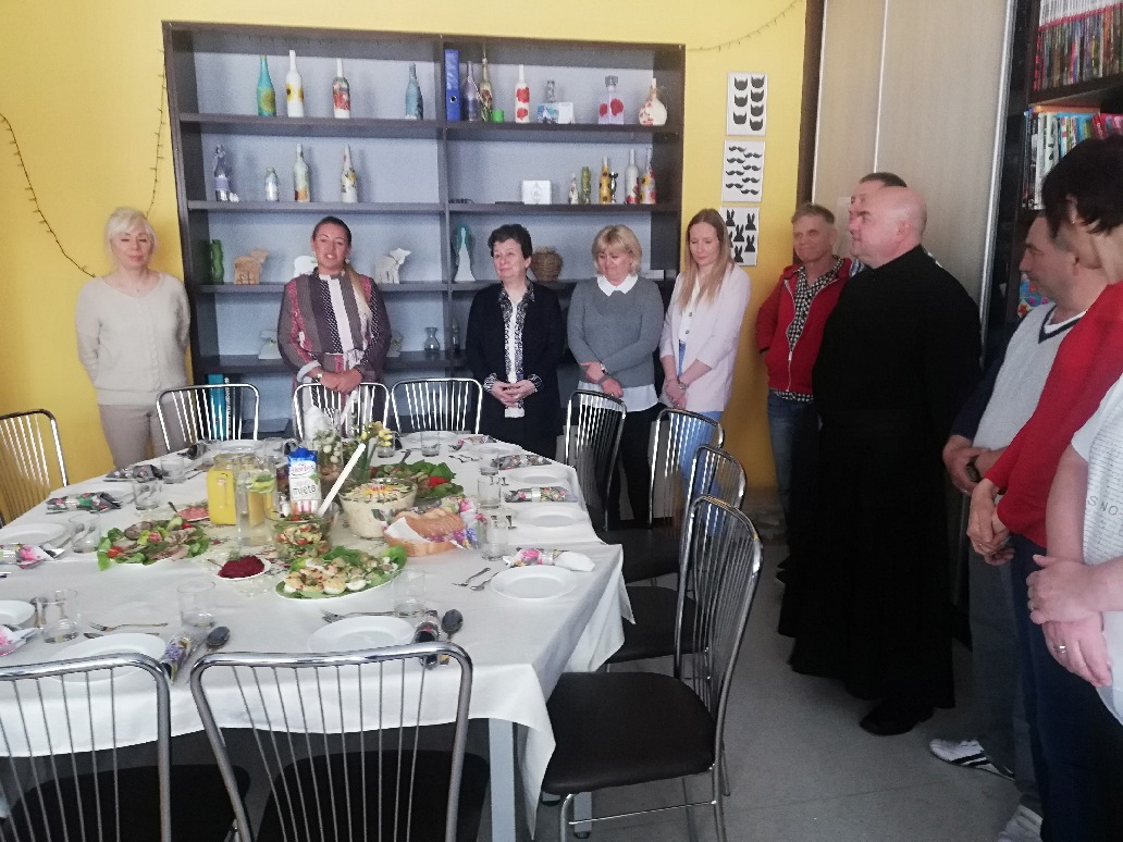 Grupa pracowników wraz z księdzem stoi w półkolu dookoła stołu. Na stole znajdują się potrawy wielkanocne. 