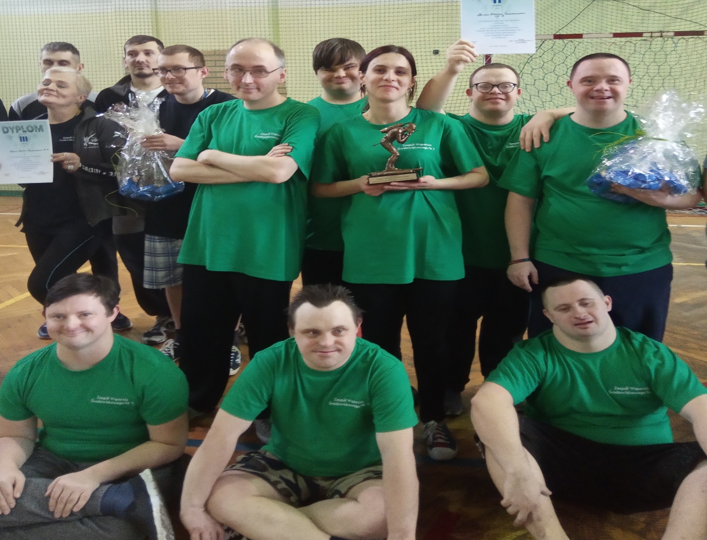 Na hali sportowej grupa osób stoi w rzędzie trzymając puchar i paczkę ze słodyczami. Przed nimi siedzi trzech uczestników. Wszyscy ubrani są w takie same koszulki. 