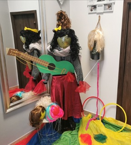 Na środku pomieszczenia stoi kukiełka z peruką, ubrana w czerwoną spódnicę marynarkę i szal z przewieszoną przez ramię tekturową gitarą. Przed i z boku postaci rozłożone są rekwizyty i akcesoria do zajęć choreograficzno - tanecznych i artystyczno – kabaretowych.
