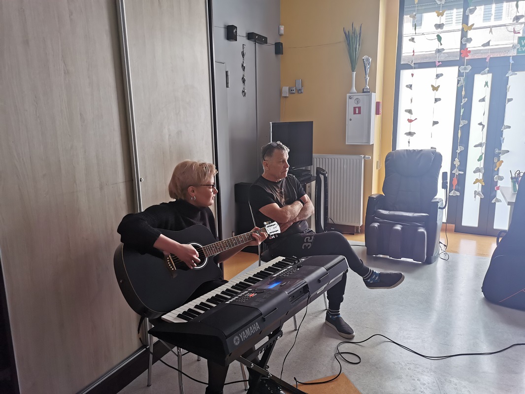 Dwoje terapeutów siedzi na krzesłach. Jeden z nich trzyma w rękach gitarę. Przed nim znajduje się keyboard. 