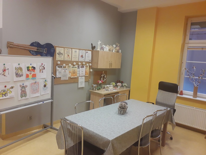 W pomieszczeniu stoi stół z krzesłami. W rogu stoi biurko, a nad nim wisi szafka na których stoją ozdoby. Na ścianie wiszą tablice z pracami plastycznymi.