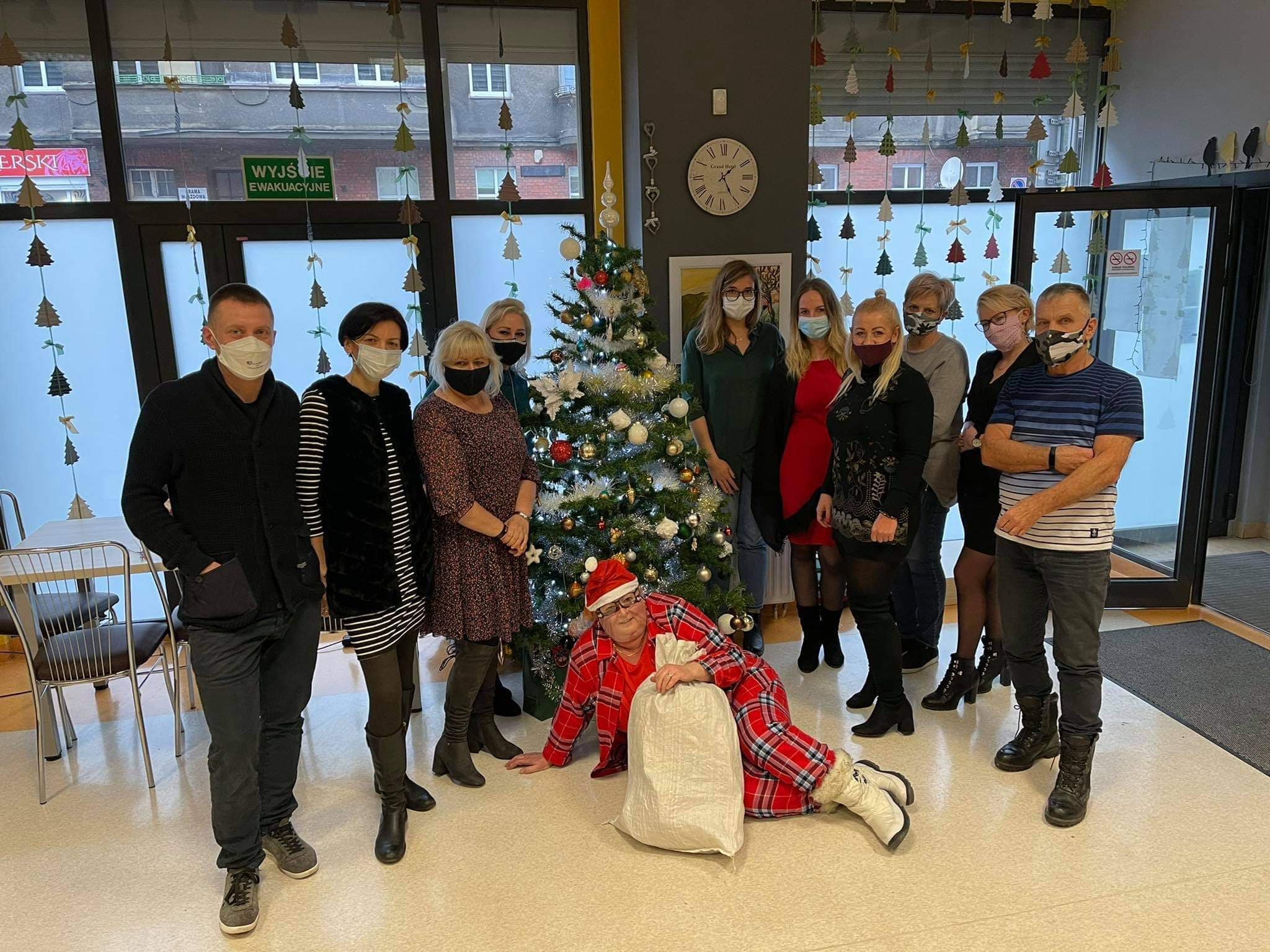 Grupa terapeutów z maseczkami na twarzach stoi w rzędzie, po dwóch stronach przystrojonej ozdobami choinki, która stoi po środku grupy. Przed nią siedzi terapeutka w przyłbicy w stroju Świętego Mikołaja, obejmując ręką worek. 