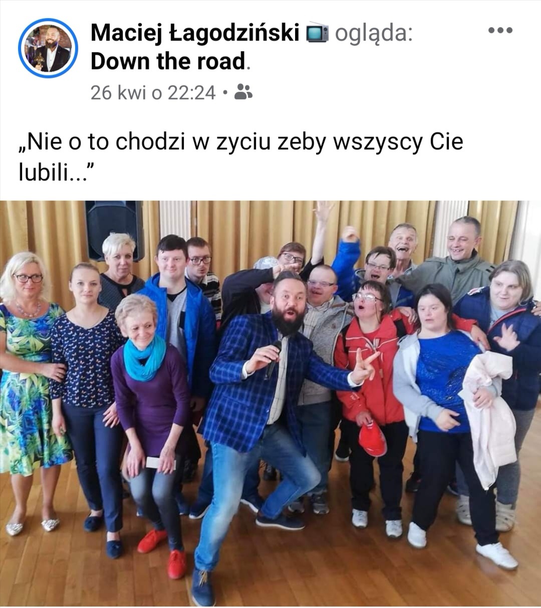 Grupa ludzi stoi na sali. Na środku grupy, z przodu stoi mężczyzna z uniesionymi rękoma do przodu. 