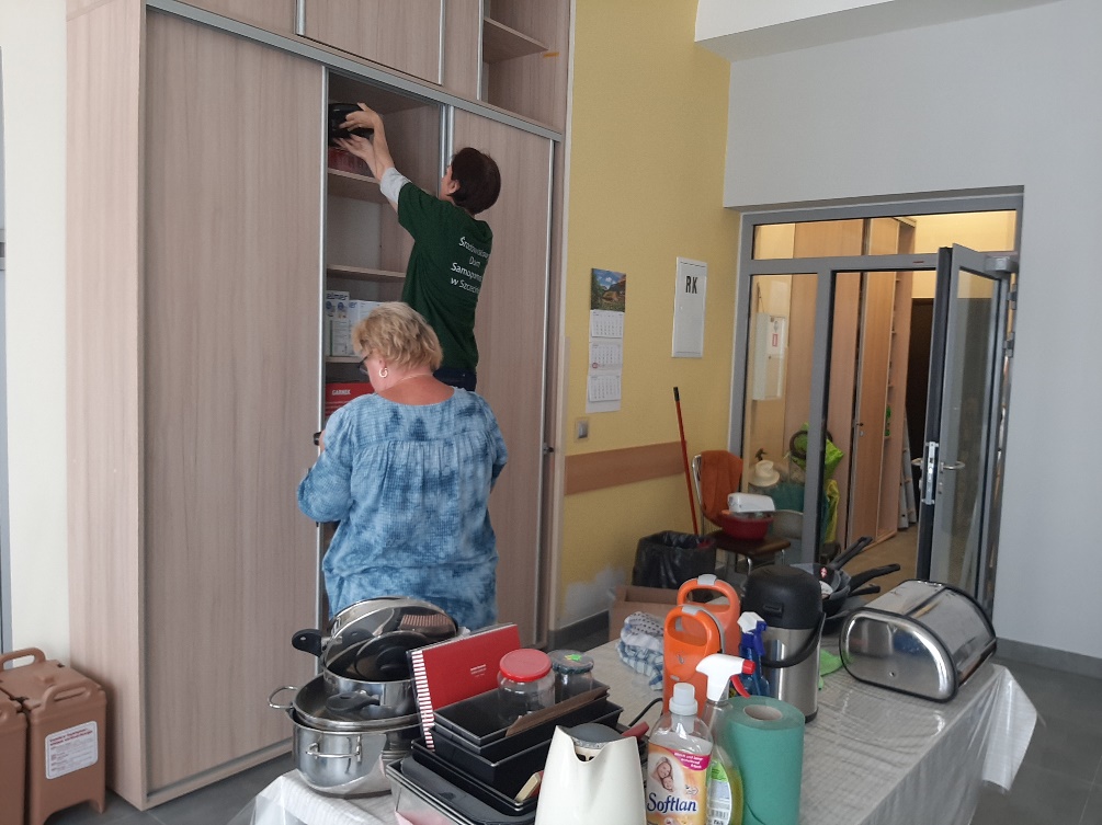 Dwie kobiety stojące tyłem. Za nimi na stole artykuły gospodarstwa domowego.