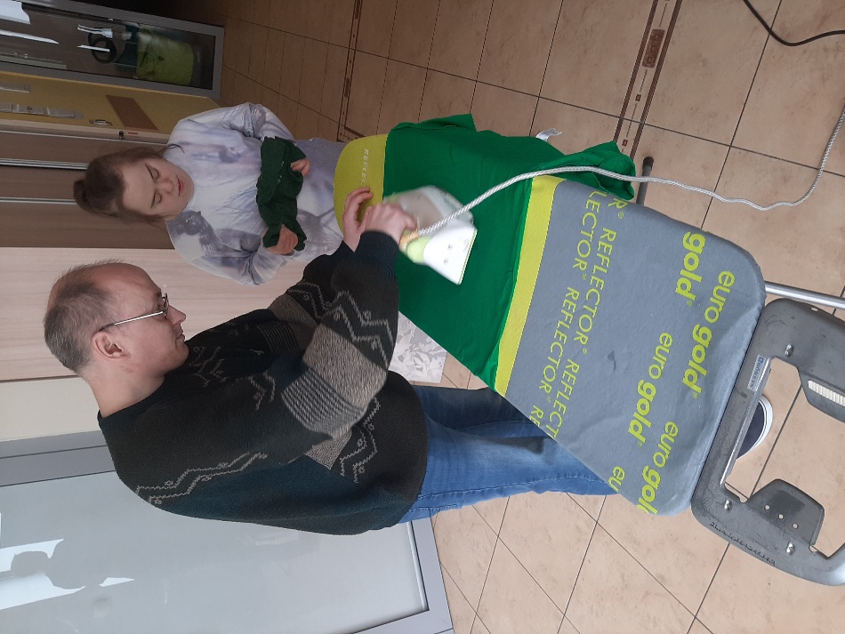 Mężczyzna prasujący żelazkiem na desce do prasowania zieloną koszulkę, obok stojąca kobieta.