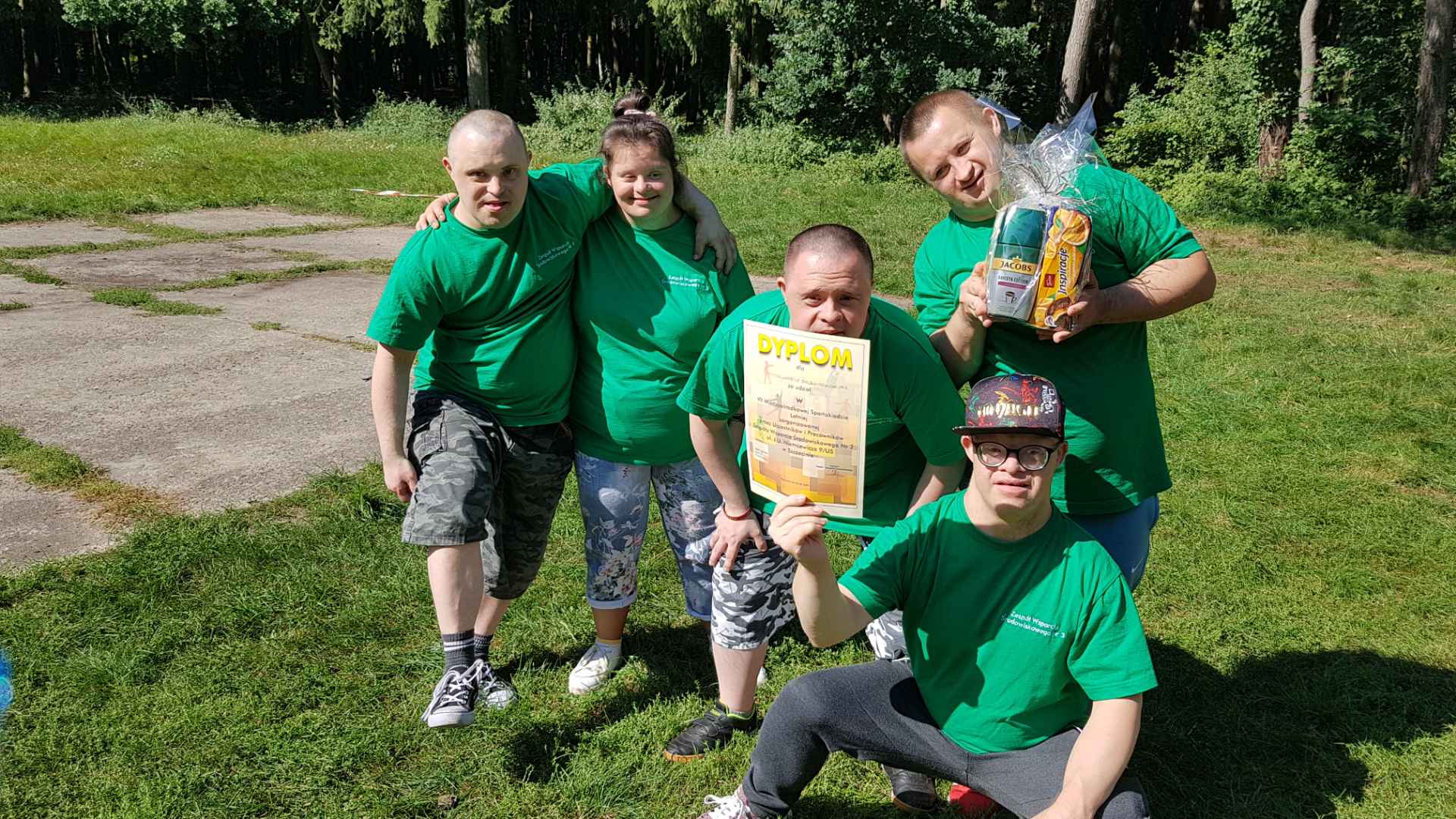 Grupa osób w zielonych koszulkach na tle lasu trzymająca w rękach dyplom. 