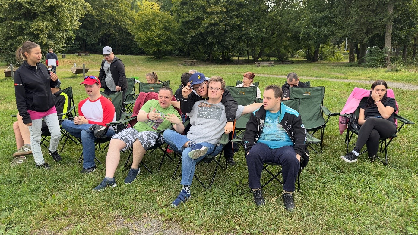 Grupa kobiet i mężczyzn siedzi na krzesłach na otwartej przestrzeni. W tle drzewa.