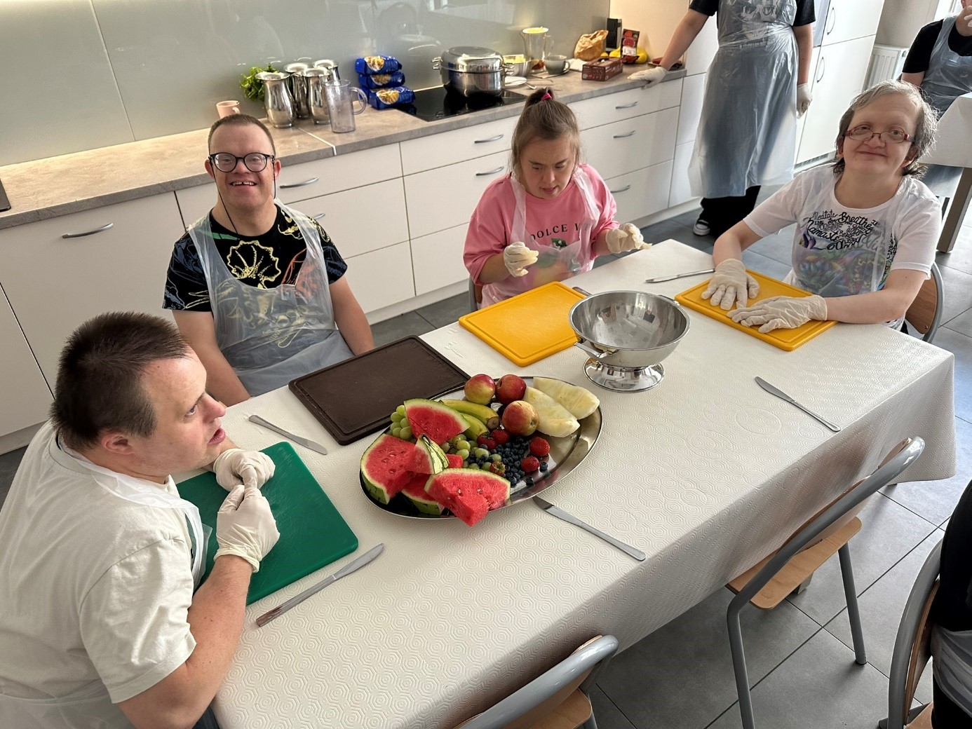 Dwóch mężczyzn i dwie kobiety siedzą przy stole. Kobiety i mężczyźni mają na sobie rękawiczki ochronne i fartuchy jednorazowe. Na stole znajdują się owoce, deski do krojenia i nóż.