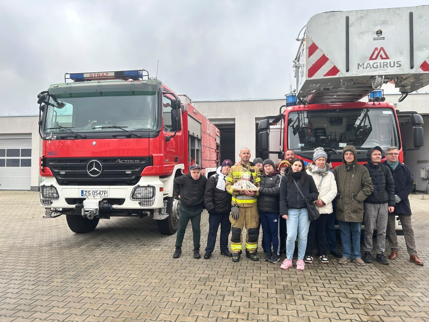 Grupa kobiet i mężczyzn pozuje do zdjęcia. W tle wozy strażackie.