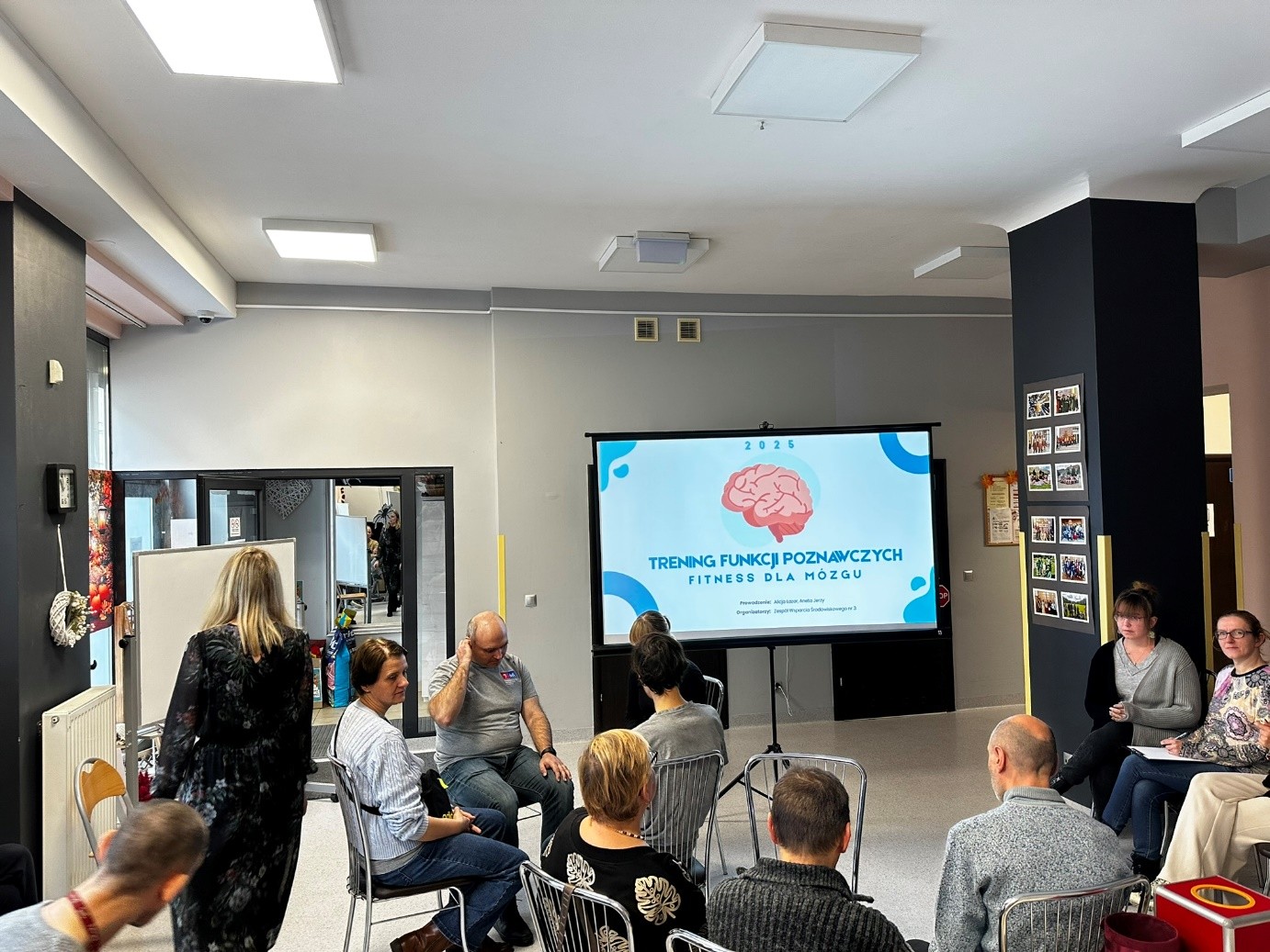 Grupa kobiet i mężczyzn siedzi na krzesłach. Na ekranie wyświetlana jest prezentacja. 
